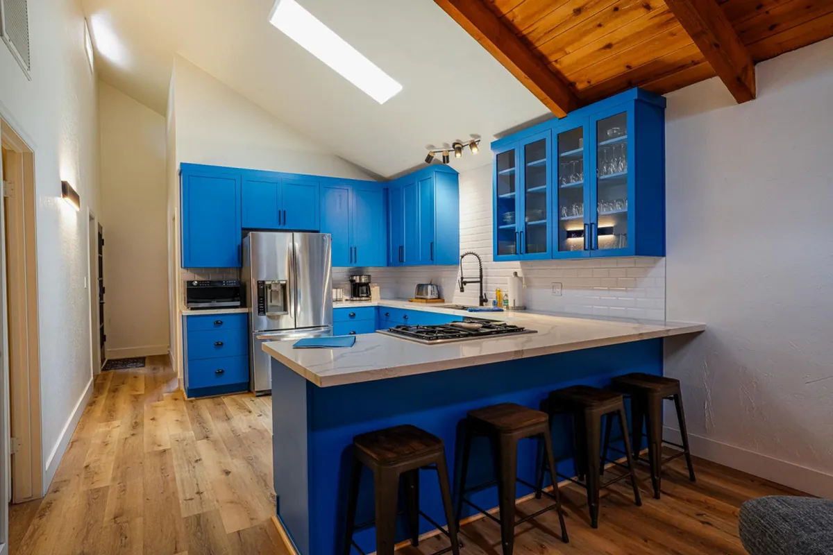 This image focuses on the kitchen island with seating, blue cabinets, and a wooden ceiling, capturing the lively and communal atmosphere. The island encourages guests to gather, cook, and converse, reinforcing the idea of everyone pitching in or lingering over food.