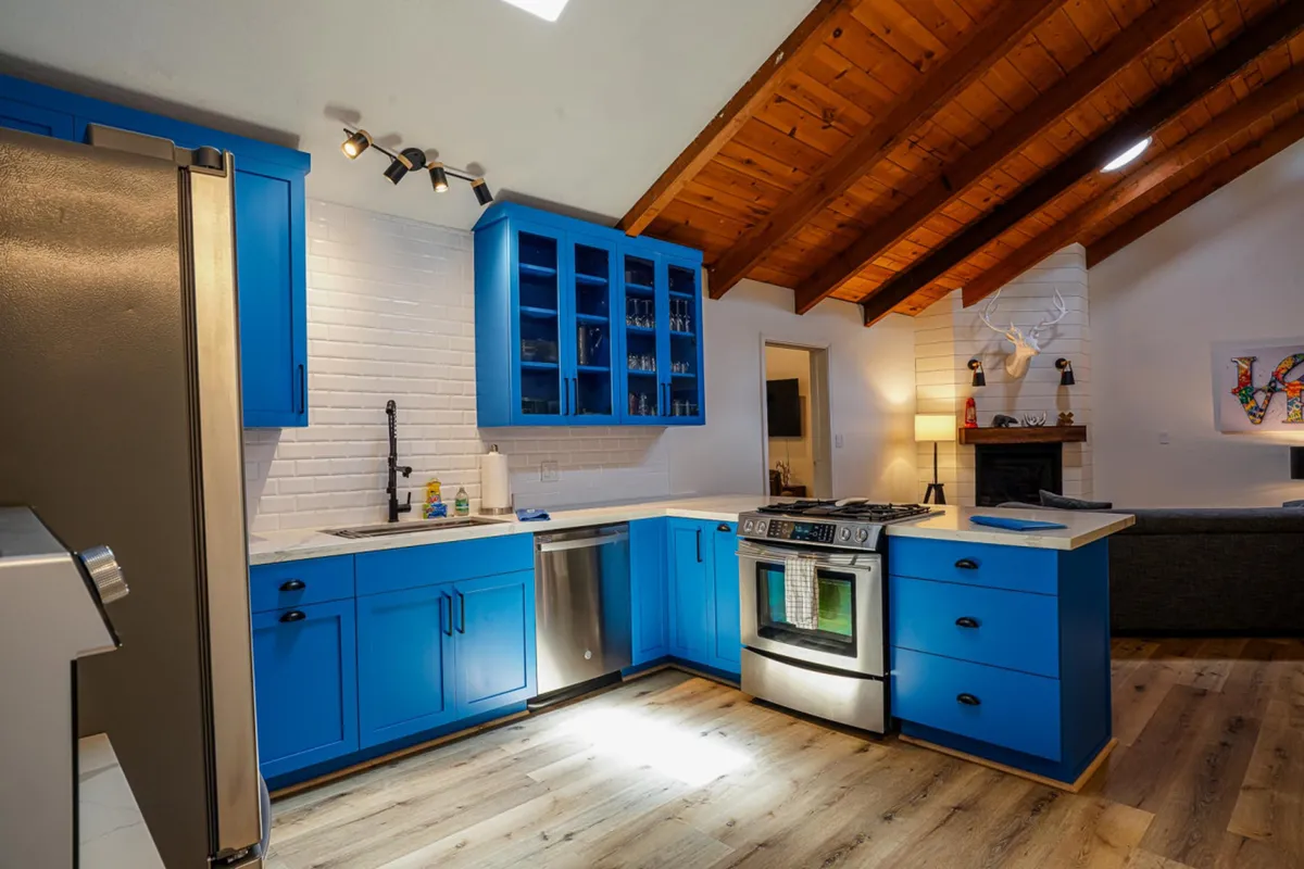 This image showcases a modern kitchen with blue cabinetry, stainless steel appliances, exposed wooden beams, and an open layout that blends seamlessly into the living space. The setting feels cheerful and welcoming, clearly designed for cooking, entertaining, and bringing people together.