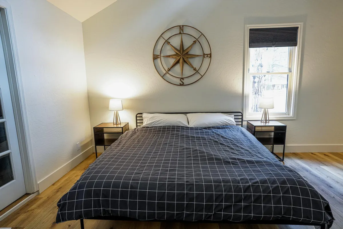 A serene bedroom with a bed draped in plaid bedding, compass wall decor, and soft natural light flowing in from a window. The space exudes peacefulness and offers a glimpse of the surrounding nature outside.