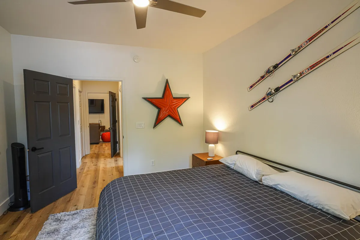 A cozy bedroom scene with a gray checkered duvet on the bed, red star wall art, skis mounted on the wall, and a nightstand with a lamp. The sunlight filtering in creates a sense of warmth and comfort, suggesting both relaxation and anticipation for outdoor adventures.