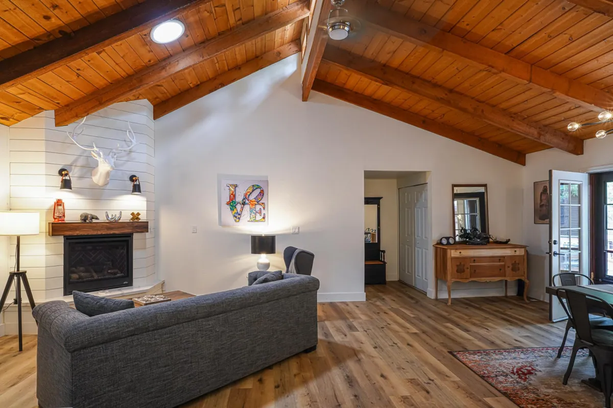 The image shows a spacious, inviting living area with a crackling fireplace as the centerpiece, surrounded by plush sofas and accented by rustic wooden beams. Artwork adorns the walls, and a generous dining table sits nearby, hinting at warm gatherings and shared meals. The overall atmosphere is cozy, perfect for relaxation and socializing.
