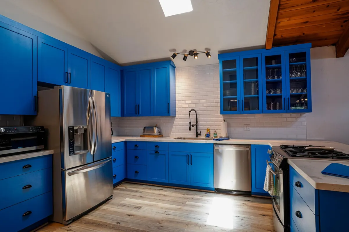 A cheerful kitchen interior featuring vibrant blue cabinets, stainless steel appliances, a white backsplash, and a warm wooden ceiling. The setup is inviting for group cooking and social gatherings, with ample space for guests to interact while preparing meals.