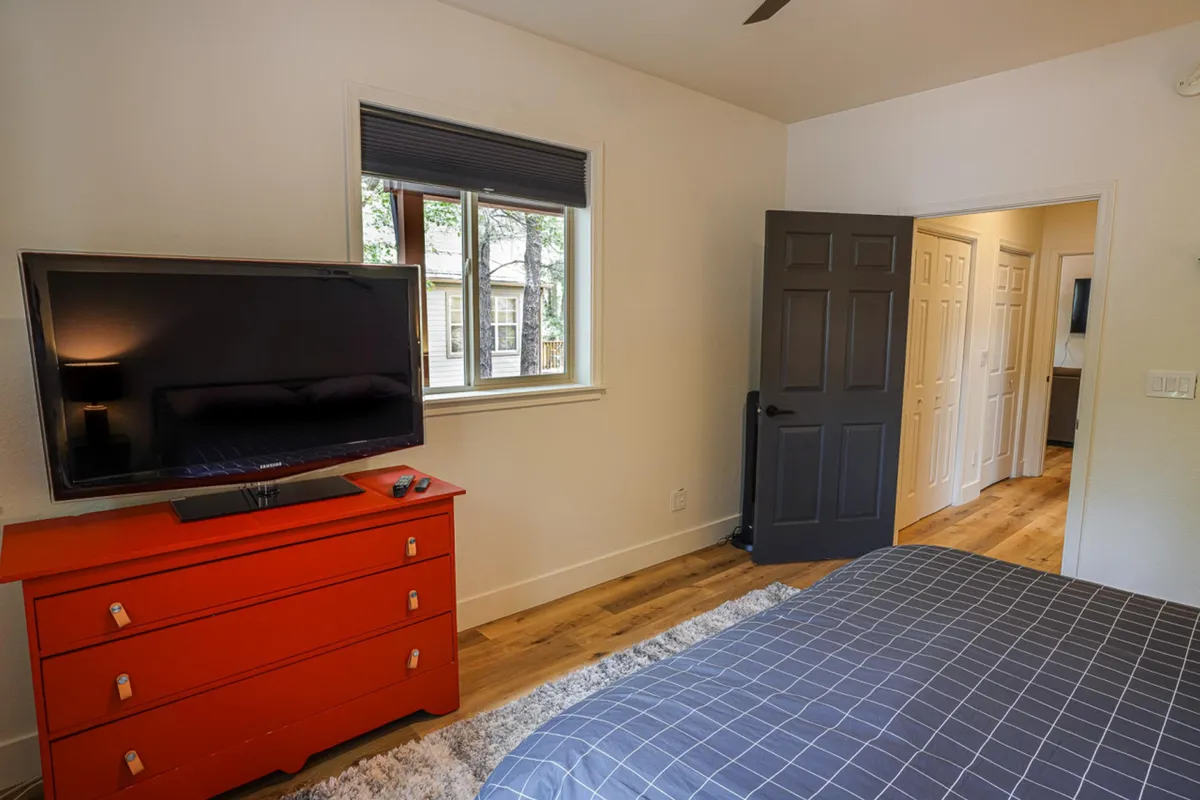 A comfortable bedroom featuring patterned bedding, a red dresser, television, and a window offering a glimpse outside. The mood is relaxing, with the window suggesting a connection to the forested location.