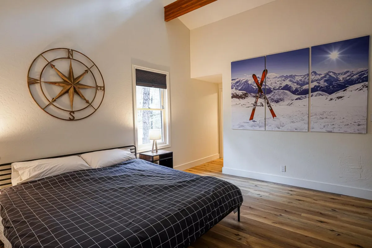 A spacious bedroom featuring plaid bedding, a large wall art of snowy mountains and skis, compass wall decor, and a sunlit window. The room exudes a calm, adventurous mood, perfect for those seeking both comfort and inspiration from the surrounding landscape.