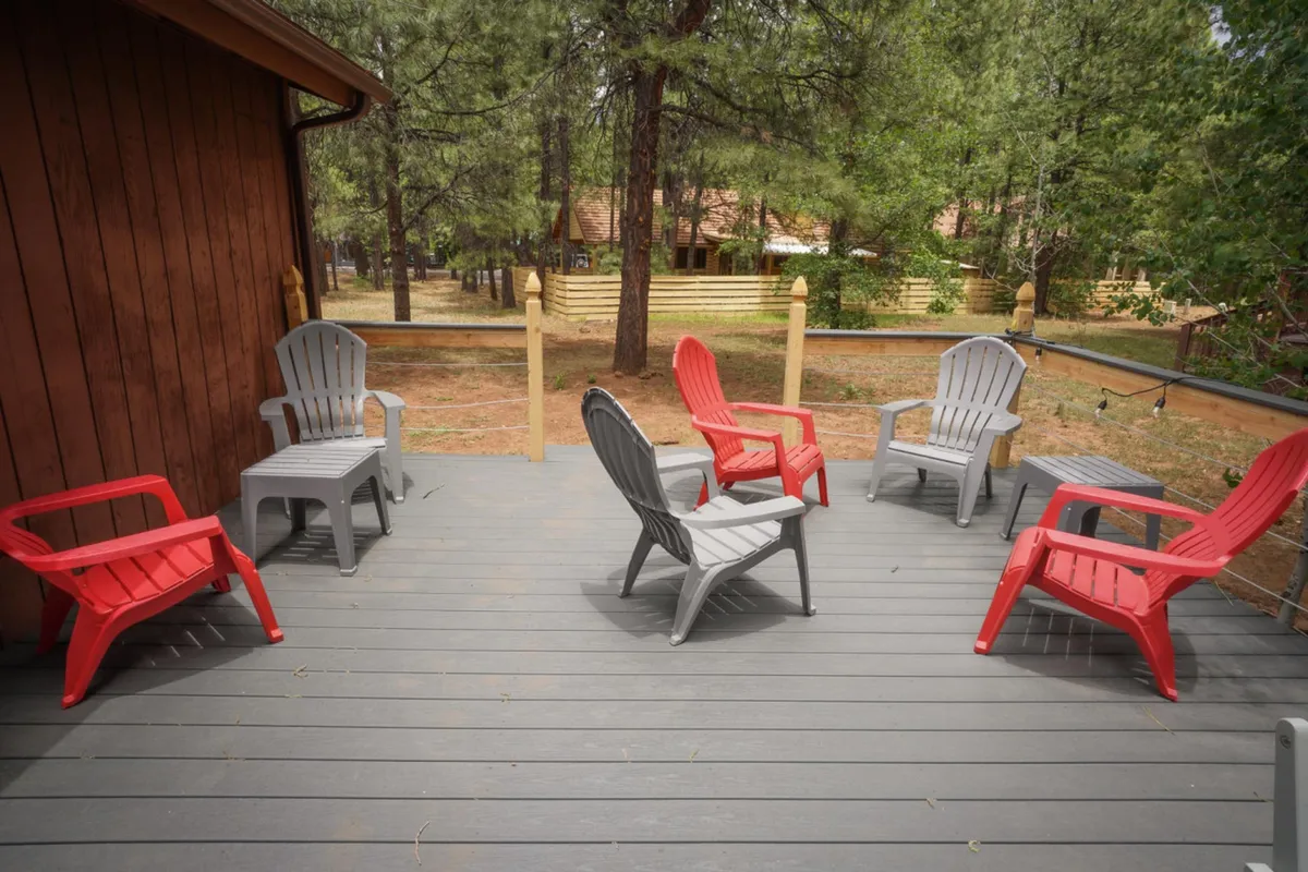 A wooden deck bordered by red and gray Adirondack chairs nestled among tall trees. The scene suggests a space for relaxing, enjoying a morning coffee, or unwinding in the fresh outdoor air, closely tied to the home’s natural surroundings.