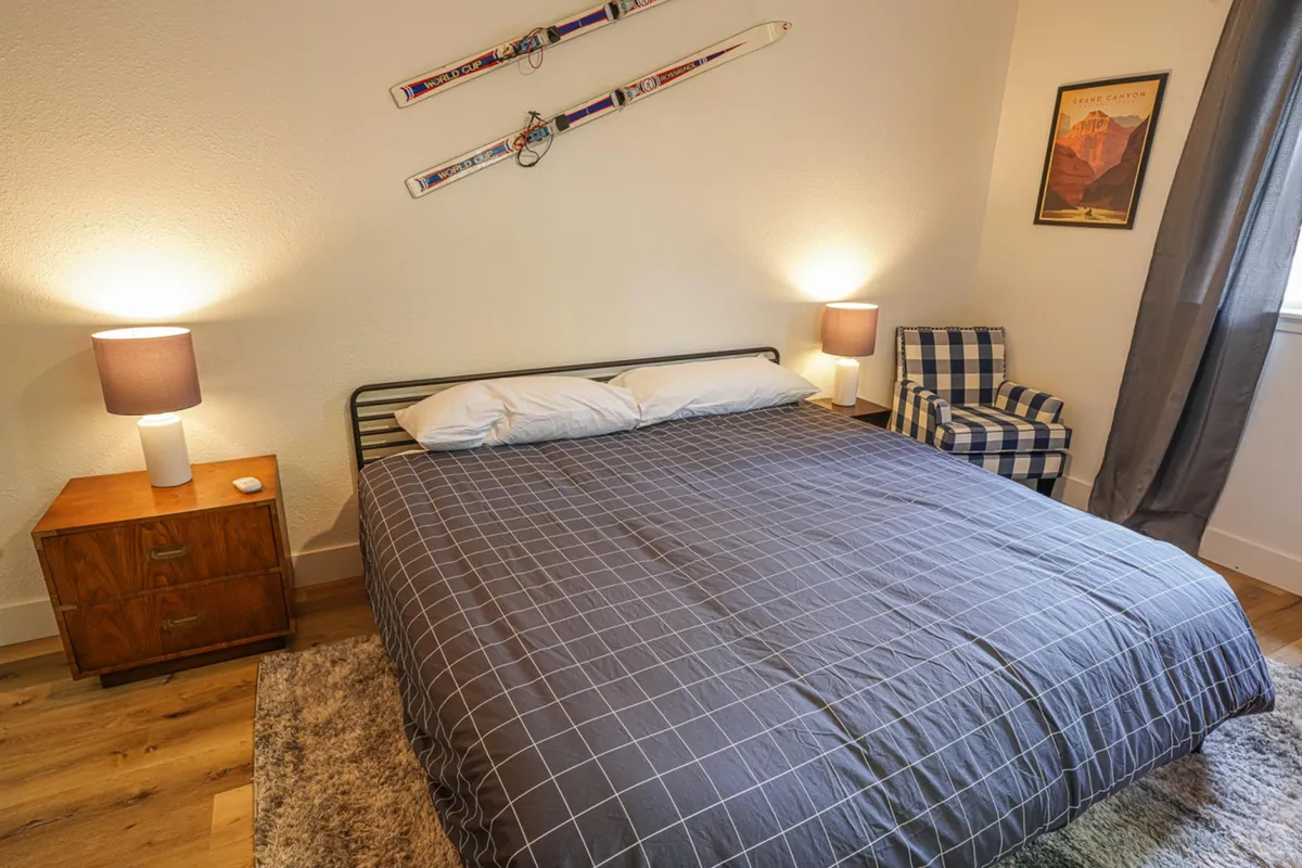 A cozy bedroom with a bed, side table and lamp, checkered chair, and ski-themed wall decor. The window lets in gentle natural light, hinting at the forested surroundings just outside, ideal for relaxing after outdoor activities.