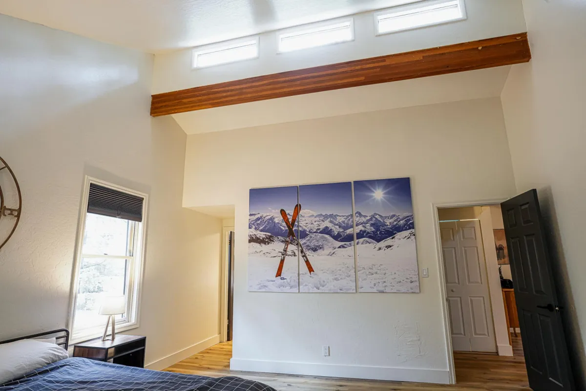 A cozy living space featuring prominent wooden beams, large artwork of skis against a snowy mountain backdrop, and abundant natural light streaming through the windows. The decor and ambiance emphasize both comfort and a connection to year-round mountain adventures.