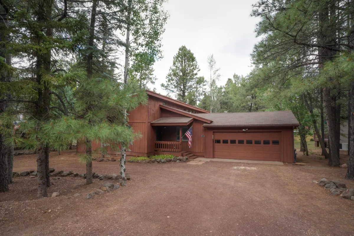 This image captures the wooden exterior of Drifting Snow Cabin set among tall pine trees, with an American flag gently waving. The gravel driveway and yard area suggest space for outdoor activities and easy access to nature trails.